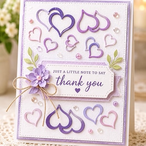 Pode incluir: Cartão de agradecimento artesanal com fundo branco texturizado e borda roxa. O cartão apresenta enfeites de coração roxos e rosa, uma flor de papel e o texto "Just a little note to say thank you".