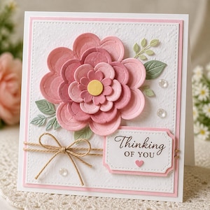 Puede incluir: Tarjeta de felicitación hecha a mano con una gran flor de papel rosa en capas como punto focal. La tarjeta presenta el texto "Thinking of You" y está adornada con hilo y pequeñas hojas. La tarjeta está sobre un fondo blanco con un borde rosa.