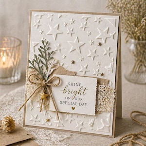 Pode incluir: Um cartão de felicitações creme com estrelas em relevo e um raminho decorativo. O cartão apresenta um rótulo quadrado com as palavras "Shine Bright on Our Special Day" e um pequeno pingente de estrela dourada.