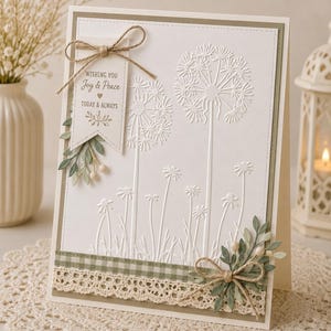 May include: A white greeting card with embossed dandelion designs. The card features a decorative banner with the text "Wishing You Joy & Peace Today & Always". Embellishments include a twine bow, greenery, and a gingham and lace border.