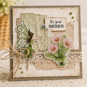 May include: Handmade birthday card featuring a fairy in a green dress with green wings, a bouquet of pink flowers, and the text "It's your Birthday." The card has a rustic, layered design with lace and twine accents.