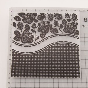 Op de afbeelding: Een doorzichtige, rechthoekige embossing folder met een rozenmotief en een geweven mandpatroon. De rozen zijn donkergrijs en zijn bovenaan gerangschikt. De folder heeft een rasterpatroon met centimeter maten.