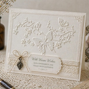 Op de afbeelding: Crèmekleurige wenskaart met een reliëf vlinder- en bloemenontwerp. Voorzien van een decoratief kanten lint, een kleine bedel en de tekst "With Warm Wishes for a Wonderful Day Filled with Happiness."