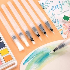 Op de afbeelding: Een verzameling aquarel penseelstiften met transparante vaten en witte borstelharen, gerangschikt op een perzik oppervlak. Een aquarelpalet en een kaart met het woord "enjoy" zijn ook zichtbaar. Ideaal voor schilderen en kalligrafie.