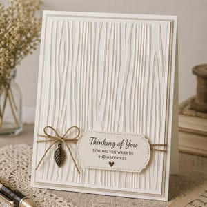 Può includere: Un biglietto di auguri color crema con una texture a venature di legno. Il biglietto presenta un'etichetta decorativa con il testo "Thinking of You" e un piccolo cuore. Un fiocco di spago e un ciondolo a forma di foglia di metallo aggiungono dettagli.