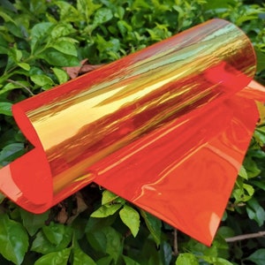 A rolled sheet of iridescent orange and gold material. The material reflects light, creating a shimmering effect. The edges of the sheet are a solid orange color. The material is resting on a bed of green leaves.
