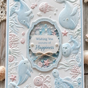May include: A rectangular greeting card featuring embossed light blue and pink sea creatures including a whale, dolphin, narwhal, octopus, and turtle. A central shaker window contains blue and white sequins with the text Wishing You oceans of Happiness.