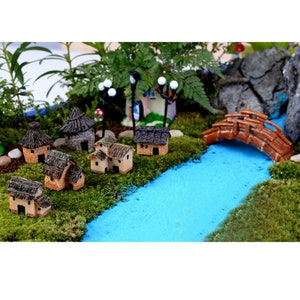 Op de afbeelding: Een miniatuur dorpscene met bruine huizen, een houten brug en een blauwe rivier. De scène is gelegen in een groen, met mos bedekt landschap met kleine bomen en een rotsformatie.