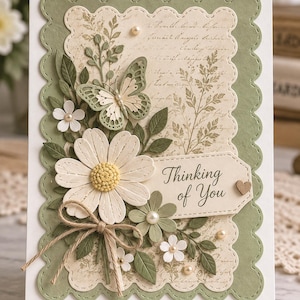 May include: A greeting card with a scalloped sage green border and a cream-coloured centre featuring vintage script text. The card is decorated with a large white daisy, a sage green butterfly, smaller white flowers, green leaves, a jute twine bow, and a tag with the text Thinking of You.