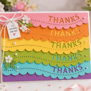 May include: A rectangular greeting card featuring five horizontal scalloped layers in pink, orange, yellow, green, and blue, each with the word THANKS die-cut into the paper. A small white tag reads SO GRATEFUL FOR YOU with a pink heart icon.