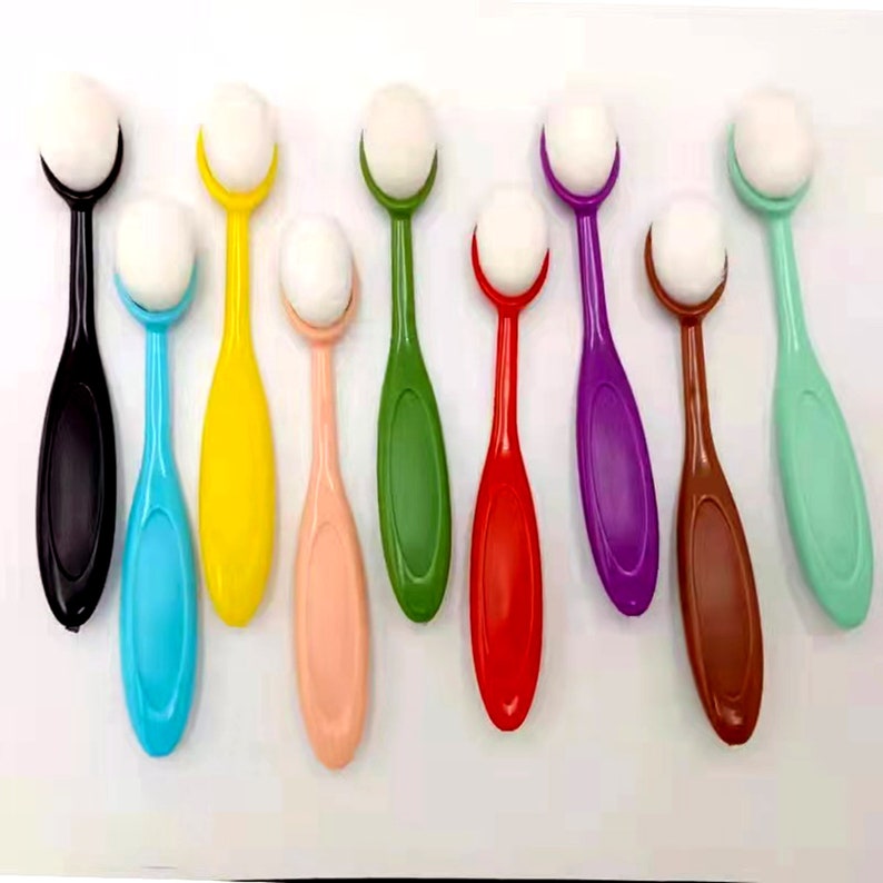 May include: An assortment of eight colorful blending brushes. Each brush features a long, oval-shaped handle in a different color, including black, blue, yellow, pink, green, red, purple, brown, and mint green. The brush heads are white and oval.