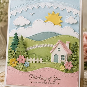 Pode incluir: Um cartão de felicitações em papel em camadas com uma paisagem que inclui um sol amarelo, nuvens brancas, colinas verdes, pinheiros, uma pequena casa com telhado rosa e flores coloridas. O texto diz Thinking of You SENDING LOVE & SMILES.