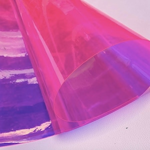A sheet of iridescent, translucent material in shades of pink and purple. The material is rolled up, revealing a layered effect of colors. The surface reflects light, creating a shimmering appearance.