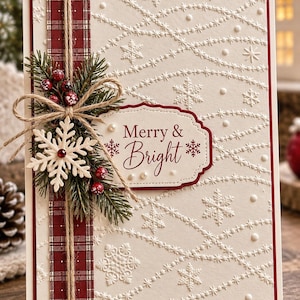 Puede incluir: Una tarjeta de felicitación color crema con copos de nieve y líneas punteadas en relieve, una cinta roja a cuadros vertical, un lazo de yute, agujas de pino verdes, bayas rojas y una etiqueta con el texto Merry & Bright en rojo.