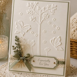 May include: A cream-colored greeting card featuring an embossed design of a tree, butterflies, and small flowers. The card is decorated with a sage green ribbon, a sprig of dried greenery, and a label with the text Especially For You.