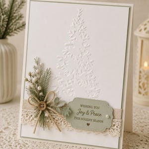 Può includere: Un biglietto di Natale bianco con un disegno ad albero in rilievo. Il biglietto presenta un rametto di verde, un nastro di juta e un'etichetta con la scritta "Wishing you Joy & Peace THIS HOLIDAY SEASON."