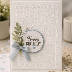 May include: A white rectangular greeting card featuring an embossed background with repeated Happy Birthday text, icons, and a central circular medallion. The medallion displays Happy Birthday in dark text with a light blue ribbon and green leaves.