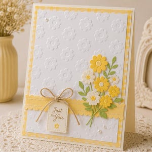 May include: A handmade greeting card with a white embossed floral pattern and a yellow gingham border. It features a bouquet of yellow and white paper flowers, a yellow ribbon, and a tag that reads "Just For You".