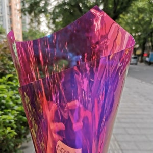 Pu&ograve; includere: Primo piano di carta cellophane iridescente multicolore. La carta &egrave; piegata, creando un effetto a strati con sfumature di rosa, viola e blu. La luce si riflette sulla superficie, creando un effetto scintillante. Lo sfondo &egrave; sfocato.