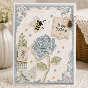 Puede incluir: Tarjeta de felicitación hecha a mano con fondo blanco texturizado y borde azul claro. La tarjeta presenta una rosa azul, una abeja, una mariposa y dos pequeñas etiquetas con las leyendas "Happy Birthday" y "Just for you". Incluye pequeños adornos azules y amarillos.