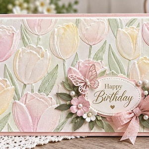 Puede incluir: Una tarjeta de felicitación de cumpleaños rectangular con un patrón en relieve de tulipanes rosa pálido y amarillos con hojas verdes. Una placa ovalada central muestra el texto Happy Birthday con un pequeño corazón, decorada con una mariposa y un lazo.