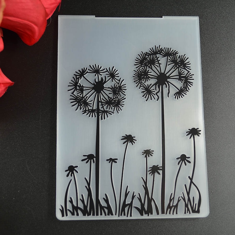 May include: A clear, rectangular embossing folder featuring a black dandelion design. Two large dandelions are centered, with smaller flowers and grass at the bottom. The folder has a slightly frosted appearance.