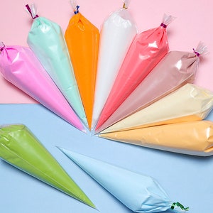 May include: A collection of colorful piping bags filled with frosting, arranged in a semi-circle. The bags contain various shades of pink, green, orange, white, and yellow. The background is split between pink and blue.