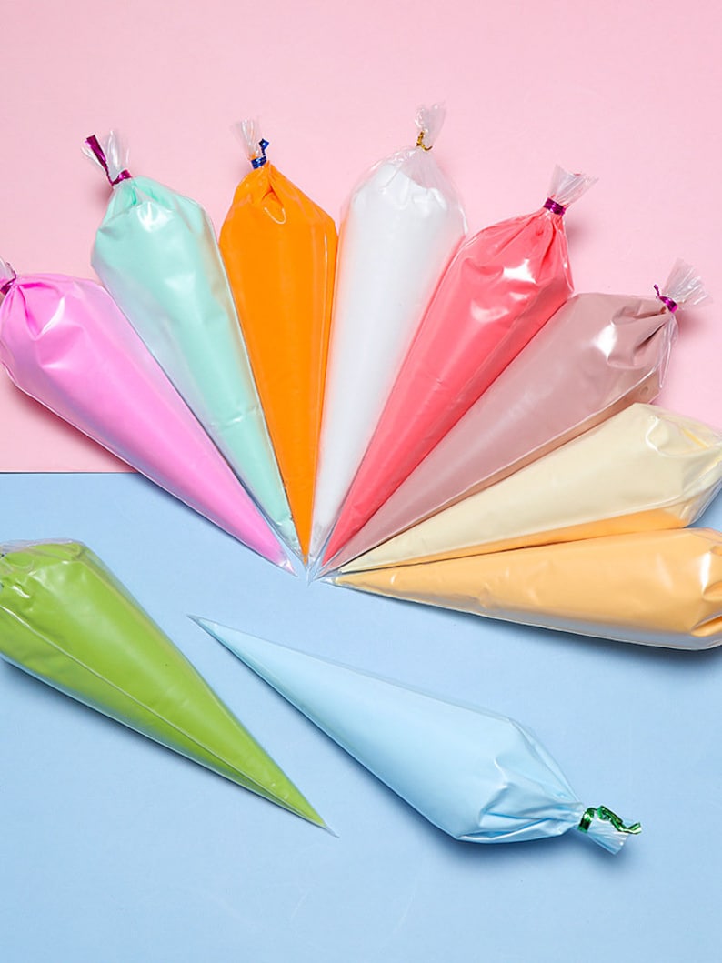 May include: A collection of colorful piping bags filled with frosting, arranged in a semi-circle. The bags contain various shades of pink, green, orange, white, and yellow. The background is split between pink and blue.