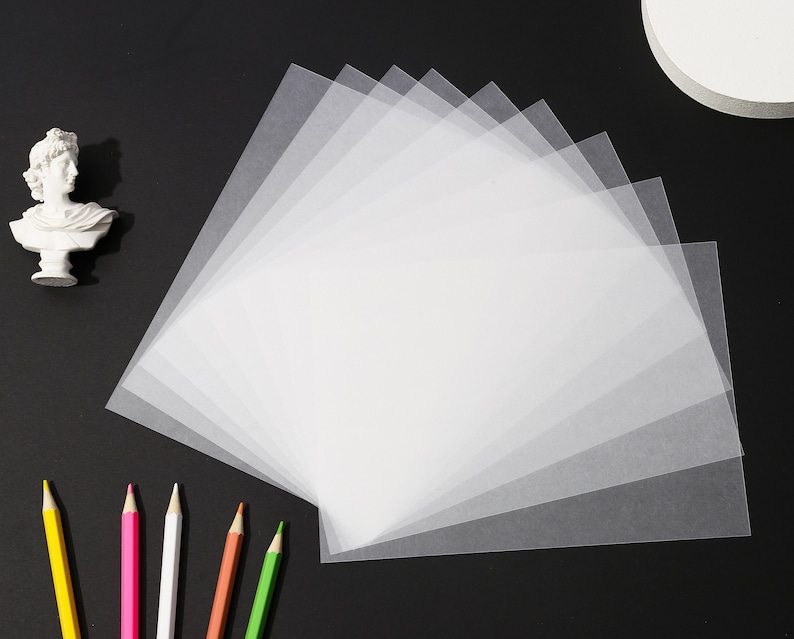 May include: A collection of translucent white sheets of paper fanned out on a black surface. A small white bust and several colored pencils are arranged around the paper. The pencils are yellow, pink, white, orange, and green.
