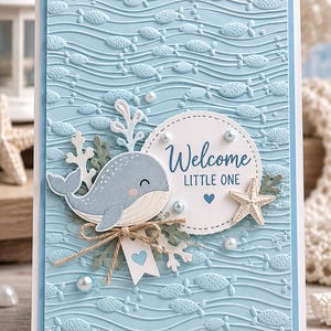 May include: A light blue greeting card featuring an embossed wavy pattern with small fish. The card displays a die-cut gray whale, a white circular tag with the text Welcome LITTLE ONE, a small starfish, twine bow, and white pearl accents.