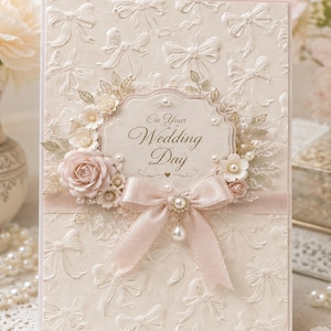 Puede incluir: Una tarjeta de felicitación de boda color crema con un patrón de lazos en relieve, una placa central festoneada con el texto On Your Wedding Day, adornada con rosas de papel rosa pálido, detalles de hojas doradas y un lazo de satén con una perla.