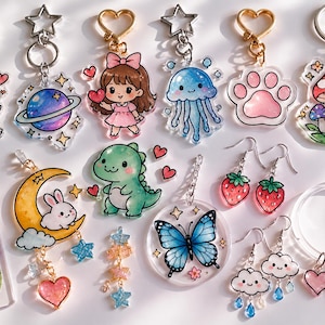 May include: A collection of colorful acrylic keychains and earrings featuring various designs. Keychains include a cat, planet, girl, jellyfish, paw print, and mushroom. Earrings showcase strawberries and clouds. A bookmark and bracelet are also visible.