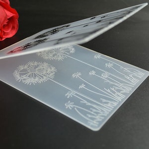 May include: A clear, rectangular, folded card with a dandelion design etched into the surface. The design features multiple dandelion seed heads and stems. The card is displayed on a dark surface.