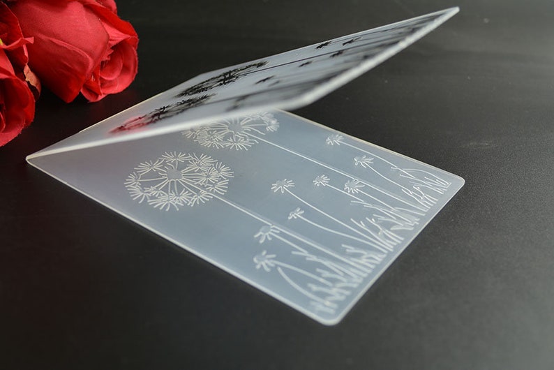 May include: A clear, rectangular, folded card with a dandelion design etched into the surface. The design features multiple dandelion seed heads and stems. The card is displayed on a dark surface.