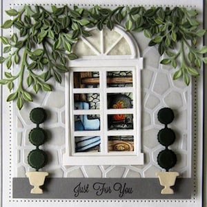 Puede incluir: Una tarjeta blanca con una ventana recortada, que muestra una escena de chimenea. La ventana está decorada con hojas verdes y dos árboles en macetas blancas. La tarjeta tiene una pancarta gris en la parte inferior con el texto "Just For You".