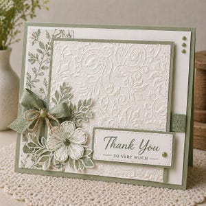 May include: A handmade thank you card with a floral design. The card features a white embossed background with a green border and accents. A green ribbon and flower embellish the front, with the words "Thank You - SO VERY MUCH" printed on a white label.
