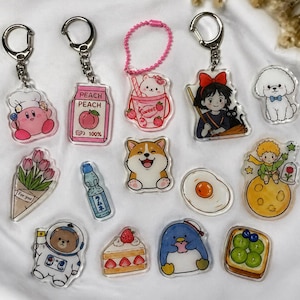 May include: A collection of clear acrylic keychains with various designs. The keychains feature cartoon characters, food items, and other cute illustrations. Some keychains have silver metal rings, while others have pink beaded chains.