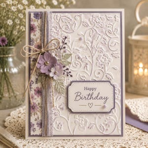 May include: A handmade birthday card with a floral design. The card features a white embossed background with a floral pattern, a purple border, and a decorative floral arrangement. The words "Happy Birthday" are written on a label with a heart symbol.