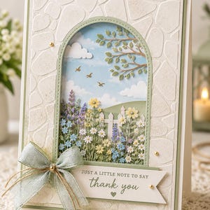 Puede incluir: Una tarjeta de felicitación hecha a mano con una escena de jardín en 3D. La tarjeta presenta un borde texturizado en forma de piedra, un lazo verde claro y el texto "gracias". La escena del jardín incluye flores, una valla blanca y un cielo azul.