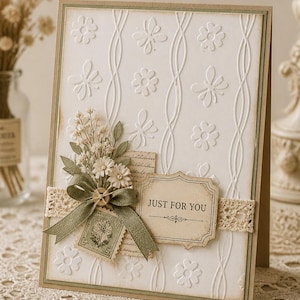 Peut inclure: Une carte de vœux faite à la main avec un motif floral en relief blanc et une bordure décorative. Elle présente un bouquet de petites fleurs, un ruban vert, un timbre-poste vintage et une étiquette indiquant "JUST FOR YOU".