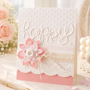 Könnte beinhalten: Eine handgefertigte Muttertagskarte mit weißem Hintergrund mit gepunkteten Prägungen und dem Wort "happy". Eine rosa Blume mit Knopfmitte und ein dekoratives Band sind angebracht. Die Karte enthält auch den Text "MOTHER'S DAY".