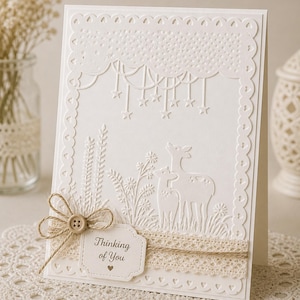 Puede incluir: Tarjeta de felicitación blanca en relieve con una escena de ciervos, estrellas y elementos florales. Presenta una etiqueta con el texto "Thinking of You" y un lazo decorativo. La tarjeta está adornada con encaje y un botón.