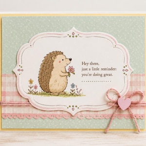 May include: A greeting card with a light green polka dot background and a pink and white plaid border. The card features a cartoon hedgehog holding a flower, with the text "Hey there, just a little reminder: you're doing great."