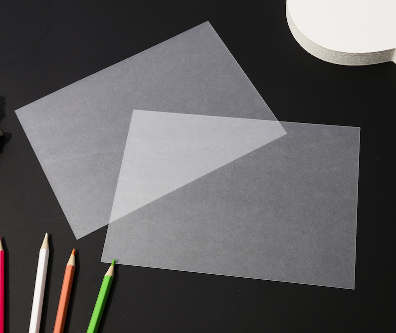 May include: Two sheets of translucent, off-white paper are arranged on a dark surface. The paper is slightly textured and appears to be thin. Colored pencils in red, white, orange, and green are visible in the lower left corner.