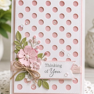 May include: A handmade greeting card with a white polka dot design over a pink background. The card features a floral arrangement with pink flowers, green leaves, and a banner that reads "Thinking of You."