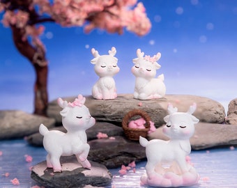 May include: Four white ceramic deer figurines with pink accents, sitting on gray rocks and a pink cloud. The deer have closed eyes and are decorated with pink flowers.
