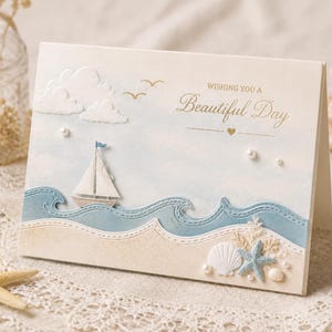 Op de afbeelding: Crèmekleurige wenskaart met een nautisch thema. De kaart toont een zeilboot op blauwe golven, een zandstrand en de tekst "Wishing You a Beautiful Day". Decoratieve elementen zijn wolken, vogels, parels, een zeester en schelpen.