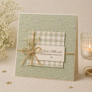 Pode incluir: Cartão de felicitações artesanal com fundo verde salvia com padrão de nuvens. Um papel xadrez é sobreposto, adornado com um laço de barbante e flores secas. O cartão diz: "Just a little note to brighten your day."