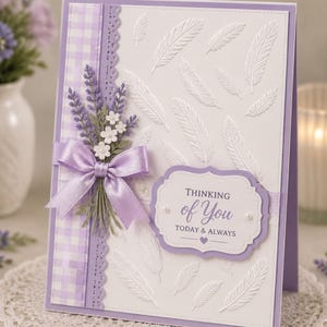 May include: A handmade greeting card with a lavender and white theme. The card features embossed white feathers on a white background. A lavender ribbon and floral arrangement adorn the left side. The card reads "Thinking of You".