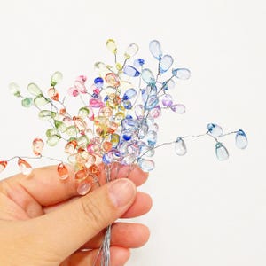 May include: A collection of colourful beaded floral stems. The teardrop-shaped beads are in shades of orange, pink, yellow, green, blue, and purple. The beads are attached to thin silver wire stems, held together in a bunch.